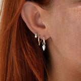 A close-up of a person's ear wearing multiple small hoop and dangle earrings, including a silver huggie hoop, a gold leaf-shaped hoop, and a silver teardrop dangle with a clear stone.